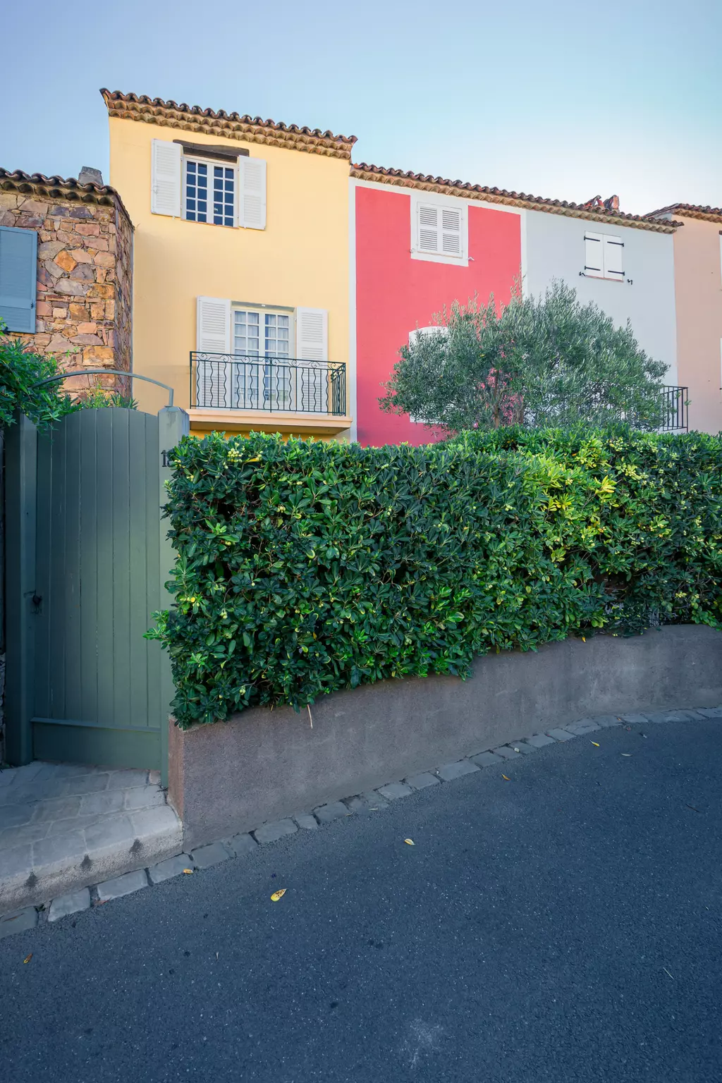 Ein schmaler Gehweg führt zu einem grünen Tor, das von einer dichten Hecke umgeben ist. Im Hintergrund sind farbige Häuser zu sehen.