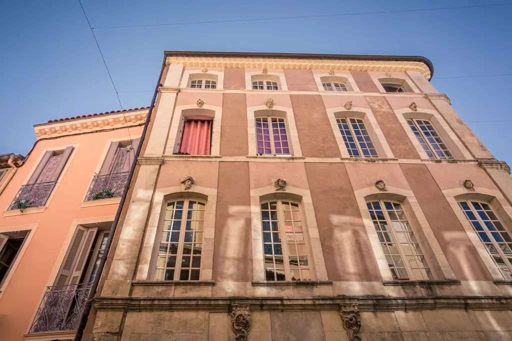 Ein mehrstöckiges Gebäude mit pastellfarbener Fassade, großen Fenstern und dekorativen Elementen. Schatten fallen auf die Wand.