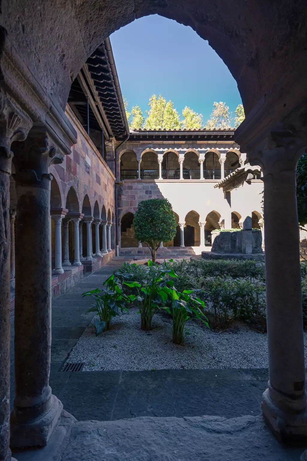 Ein Blick durch einen gotischen Bogen auf einen ruhigen Innenhof mit einem kleinen Garten und Säulen entlang der Wände.