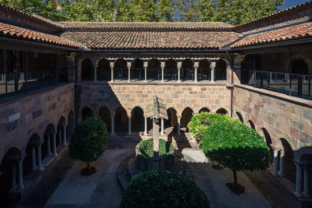 Ein Innenhof mit romanischen Bögen, Gräsern und runden, gepflegten Sträuchern um einen zentralen Brunnen.