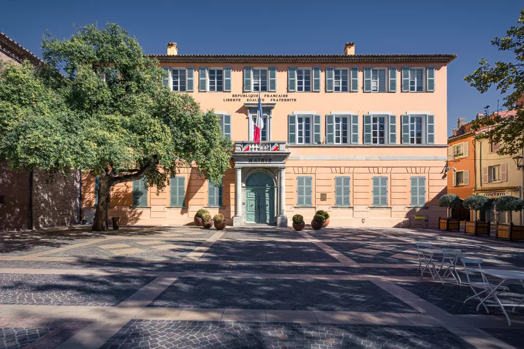 Ein historisches Gebäude mit blauen Fensterläden und einer grünen Tür, umgeben von Bäumen und einem gepflasterten Platz.