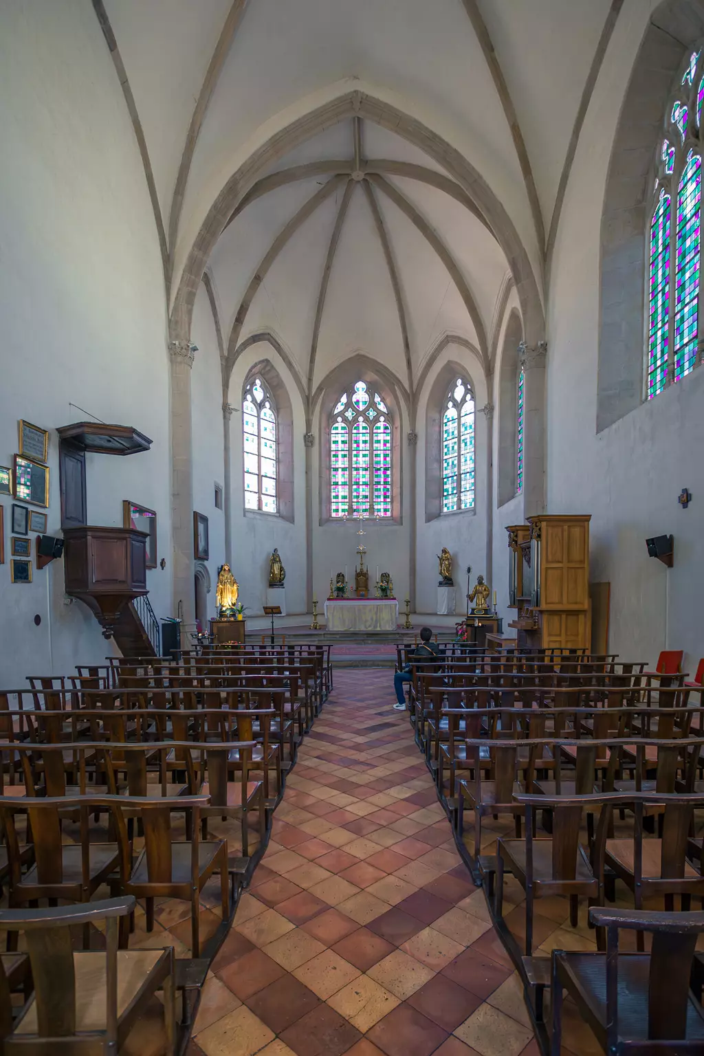 Helle Kirche mit hohen, gewölbten Decken, Holzstühlen in Reihen, bunten Fenstern und einem Altar mit Statuen im Hintergrund.