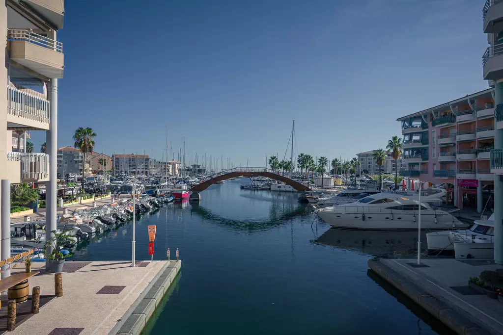 Altes Gebäude, Yachten im Wasser, eine Holzbrücke führt über den Kanal, Palmen und moderne Wohngebäude umgeben den Hafen.