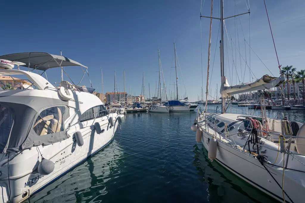 Weißes Boot im Vordergrund sowie mehrere Segelboote im Hafen, ruhiges Wasser und blauer Himmel ohne Wolken.