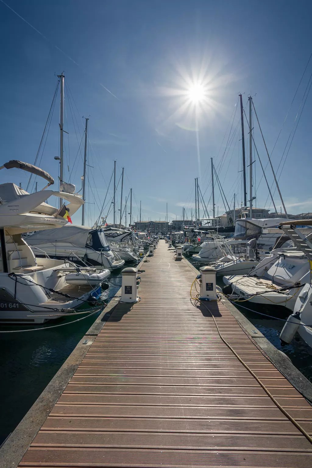 Eine Holzpier erstreckt sich zwischen Yachten im Hafen, darunter strahlt die Sonne am klaren Himmel.