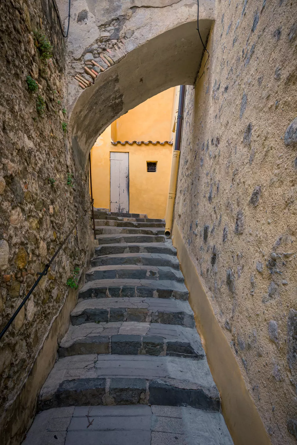 Stone staircase in a narrow alley, flanked by old walls, leading to a light yellow wall and a wooden door.