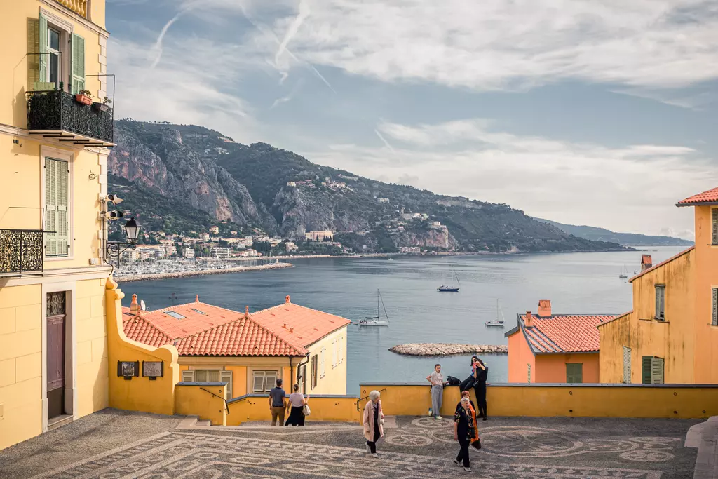 Colorful buildings and a coastal view with boats in the water, people walking on a patterned stone staircase.