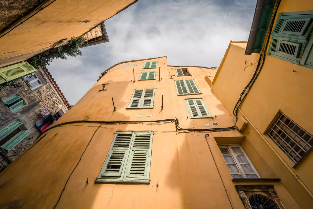 A narrow alley featuring colorful buildings with green shutters reaching up towards the sky, captured from below.