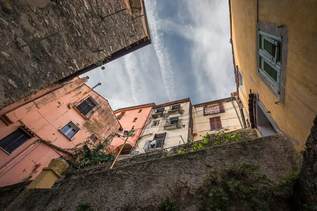 Menton: View looking up between colorful houses and city walls with a cloudy sky in the background.