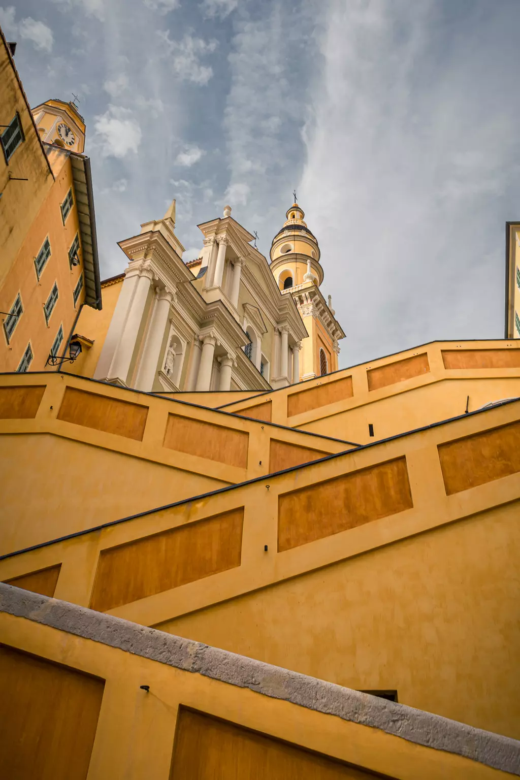 Menton: Staircases with yellow walls lead up to an ornate building topped with a tall dome and a view of the sky.