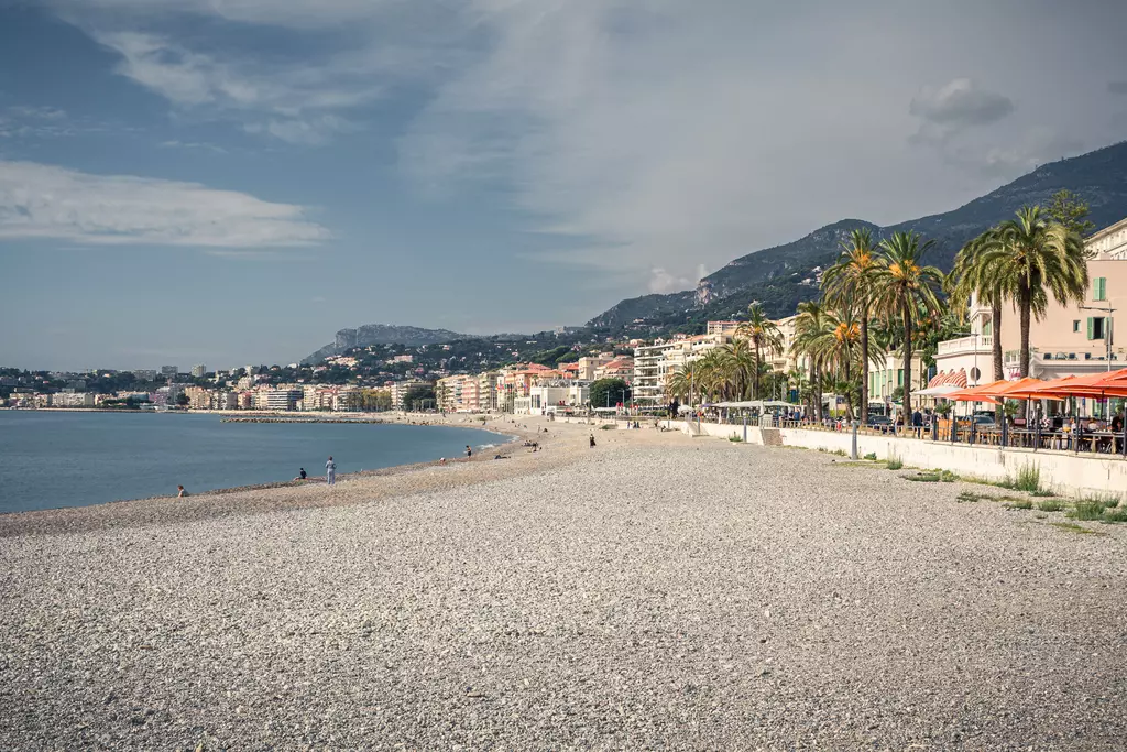 A coastal view featuring a pebble beach, surrounded by palm trees, and a glimpse of a bustling promenade with buildings.