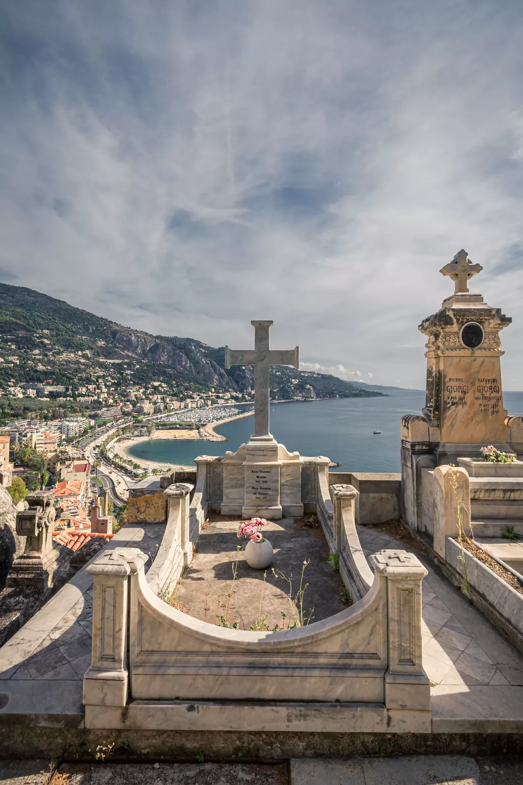 Cemetery in Menton: A cross on a mausoleum-like grave overlooking the coast and the sea, surrounded by green hills.