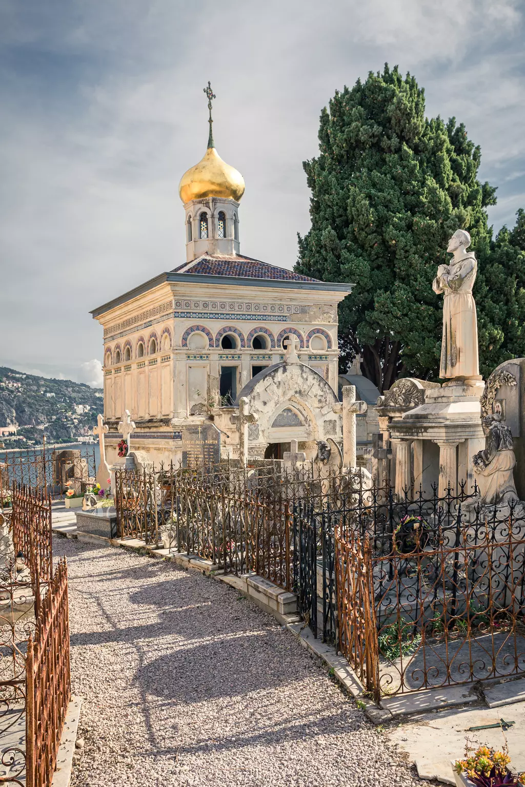 A paved path winds through a cemetery featuring a golden-domed chapel and various graves, framed by green trees.