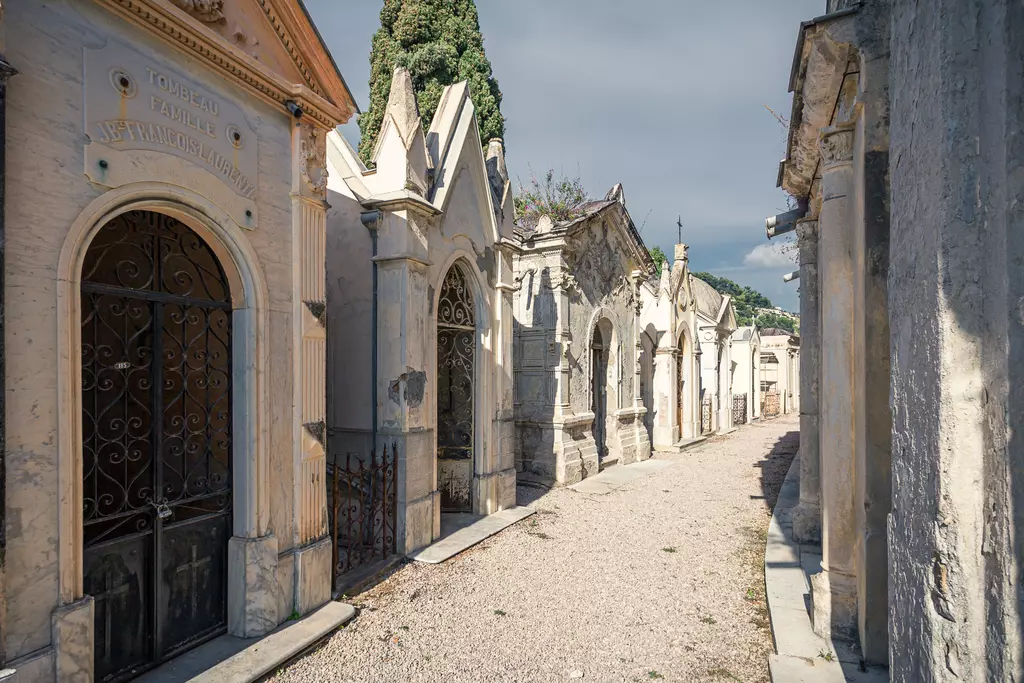 A narrow path between old, ornate tombs and mausoleums in a serene, sunlit cemetery.