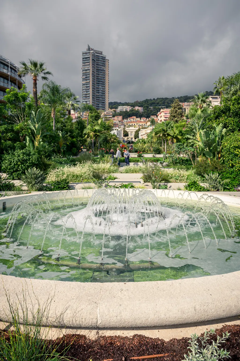 A fountain bursts with water in vibrant greenery, with modern buildings and a hilly city skyline in the background.