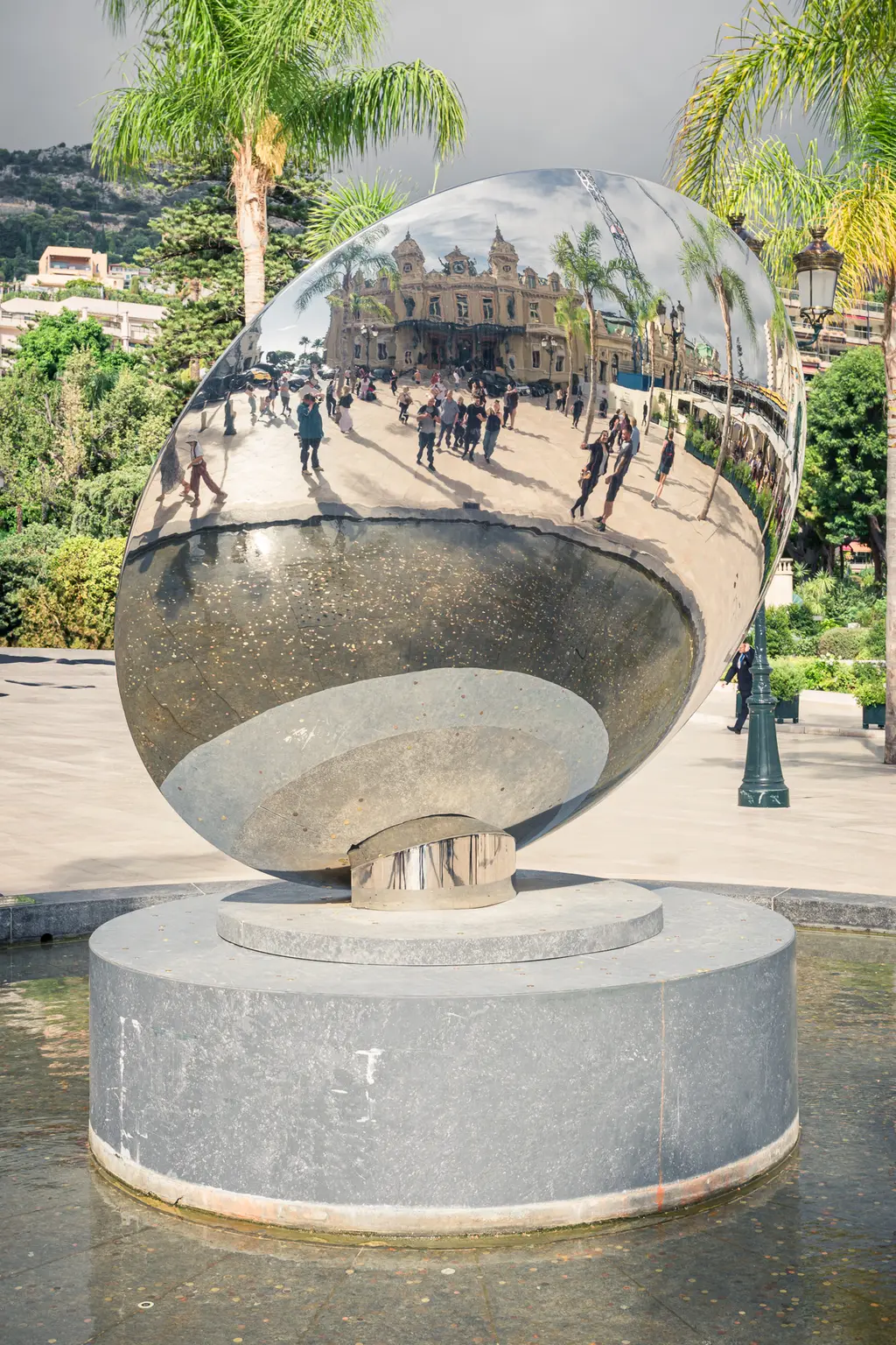 A large, shiny oval sculpture stands on a round base, surrounded by people and green palm trees.