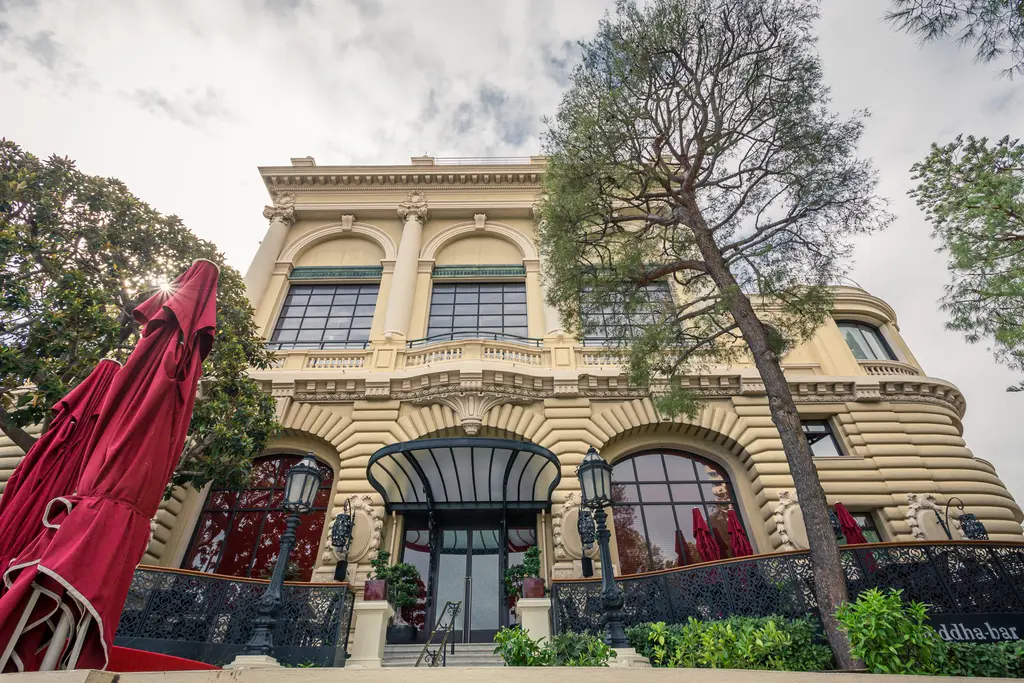 A large, elegant building view featuring a stylish facade, surrounded by trees and a red sunshade in the foreground.