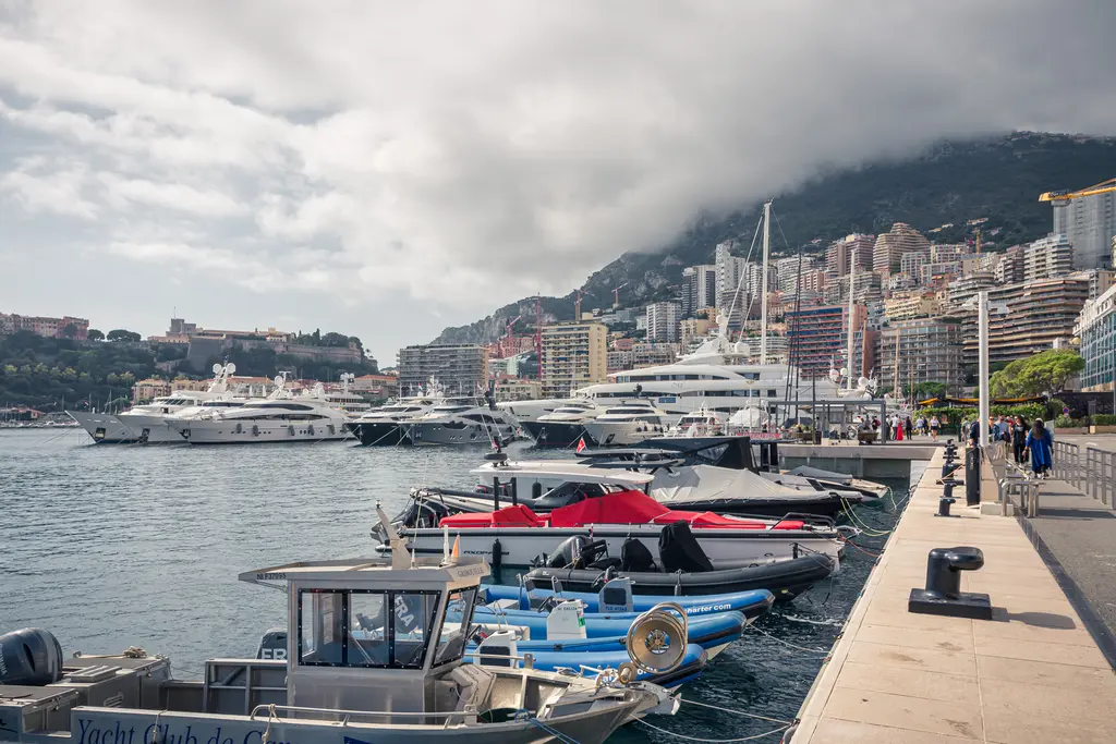 A scenic waterfront promenade with several yachts and small boats, surrounded by modern buildings and a mountain in the background.