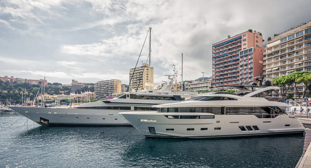 Two large yachts are moored in the harbor, surrounded by modern buildings and trees under a cloudy sky.