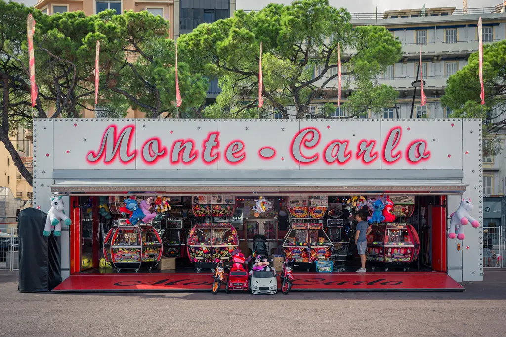A game stall with a pink sign reading Monte Carlo, surrounded by arcade machines and colorful plush toys, including two toy cars.