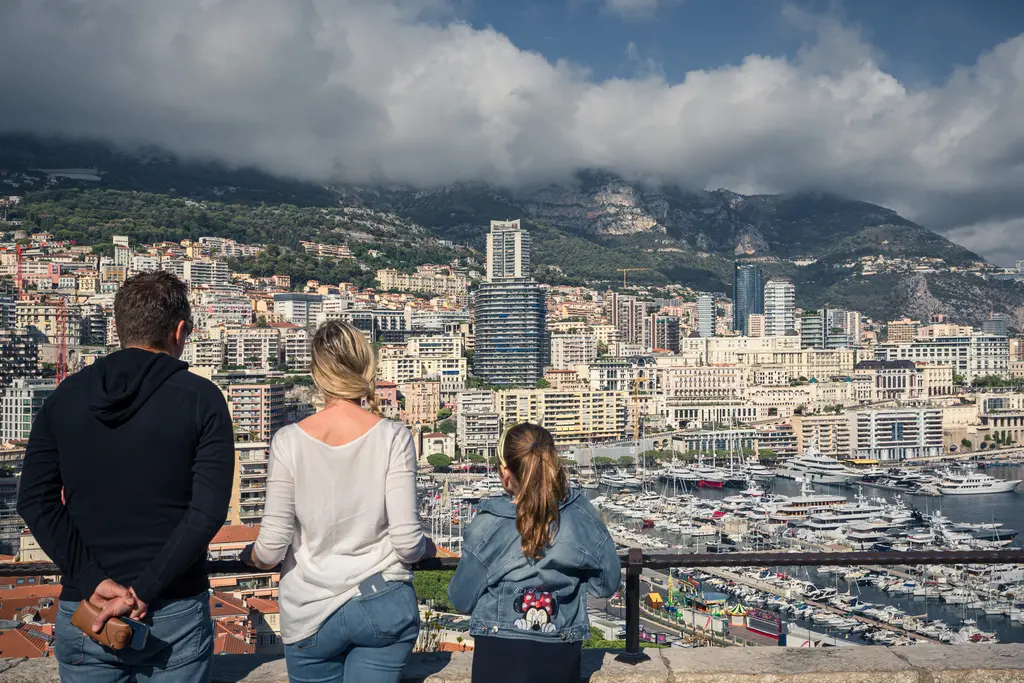 Three people are gazing at the city with modern buildings and yachts in the harbor, framed by mountains in the background.