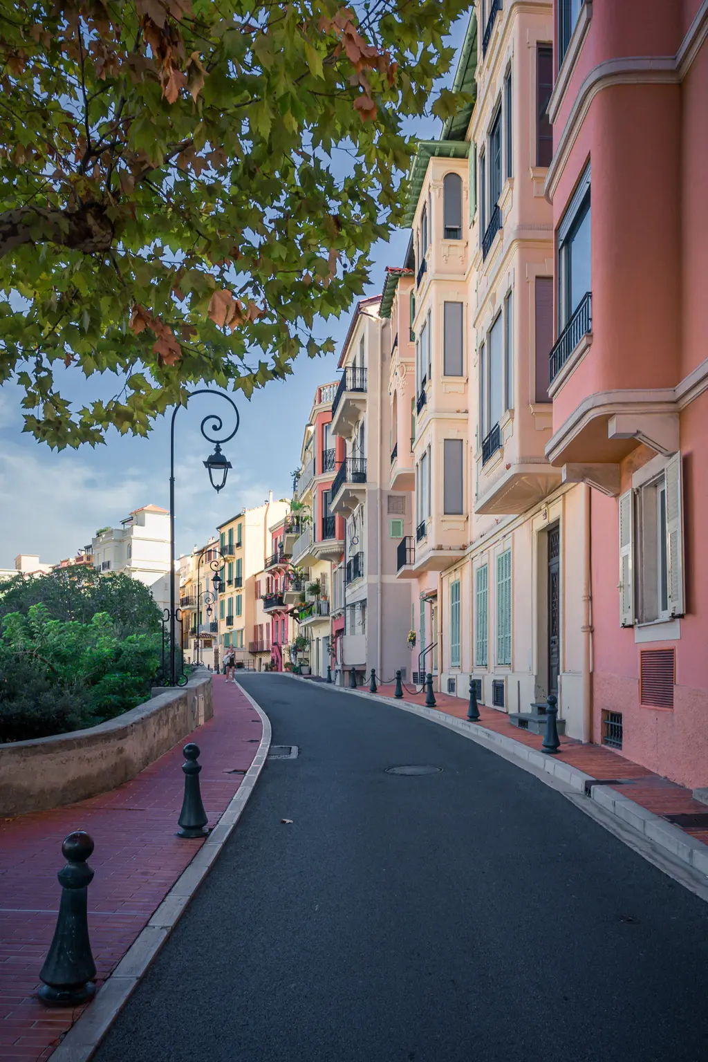 A winding street lined with colorful buildings, surrounded by trees and lampposts, stretches under a clear sky.