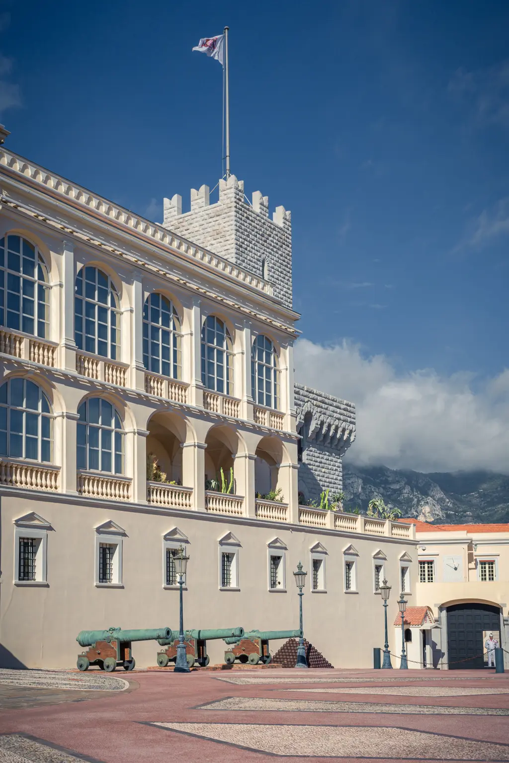 View of a historic building with tall windows and a tower, alongside old cannons on a square.