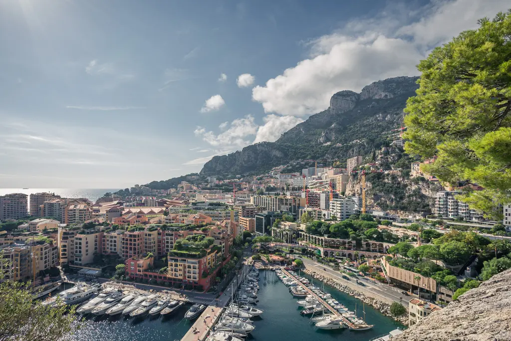 View of the harbor with yachts, surrounded by colorful buildings and mountains under a cloudy sky.