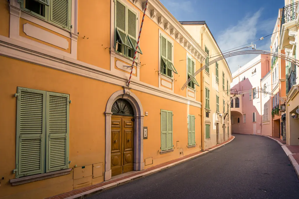 A narrow, quiet street lined with light yellow buildings featuring green shutters and a wooden door.