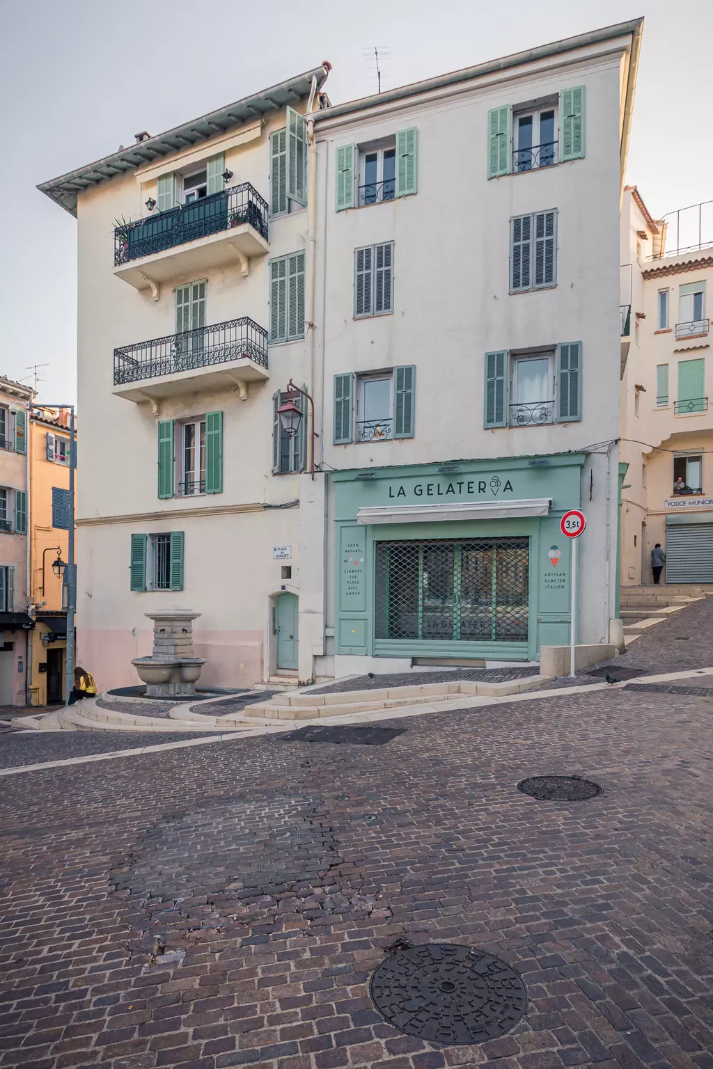 Ein helles Gebäude mit grünen Fensterläden und einer Eisdiele namens La Gelateria. Die Straße ist gepflastert und leicht ansteigend.