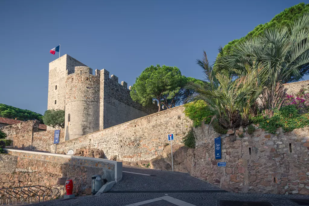 Eine Mauer aus Steinen umgibt einen alten Turm, auf dem eine französische Flagge weht. Grüne Bäume und Blumen sind sichtbar.