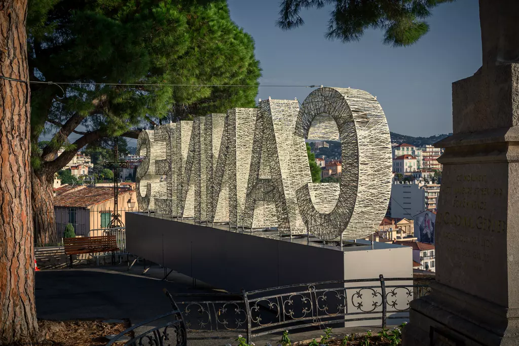 Große, verwobene Buchstaben formen das Wort Cannes, umgeben von Bäumen und mit einer Stadtansicht im Hintergrund.