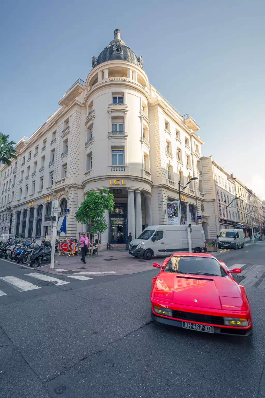 Ein rotes Sportauto fährt auf einer Straße vorbei, während ein elegantes Gebäude im Hintergrund zu sehen ist. Motorräder parken seitlich.