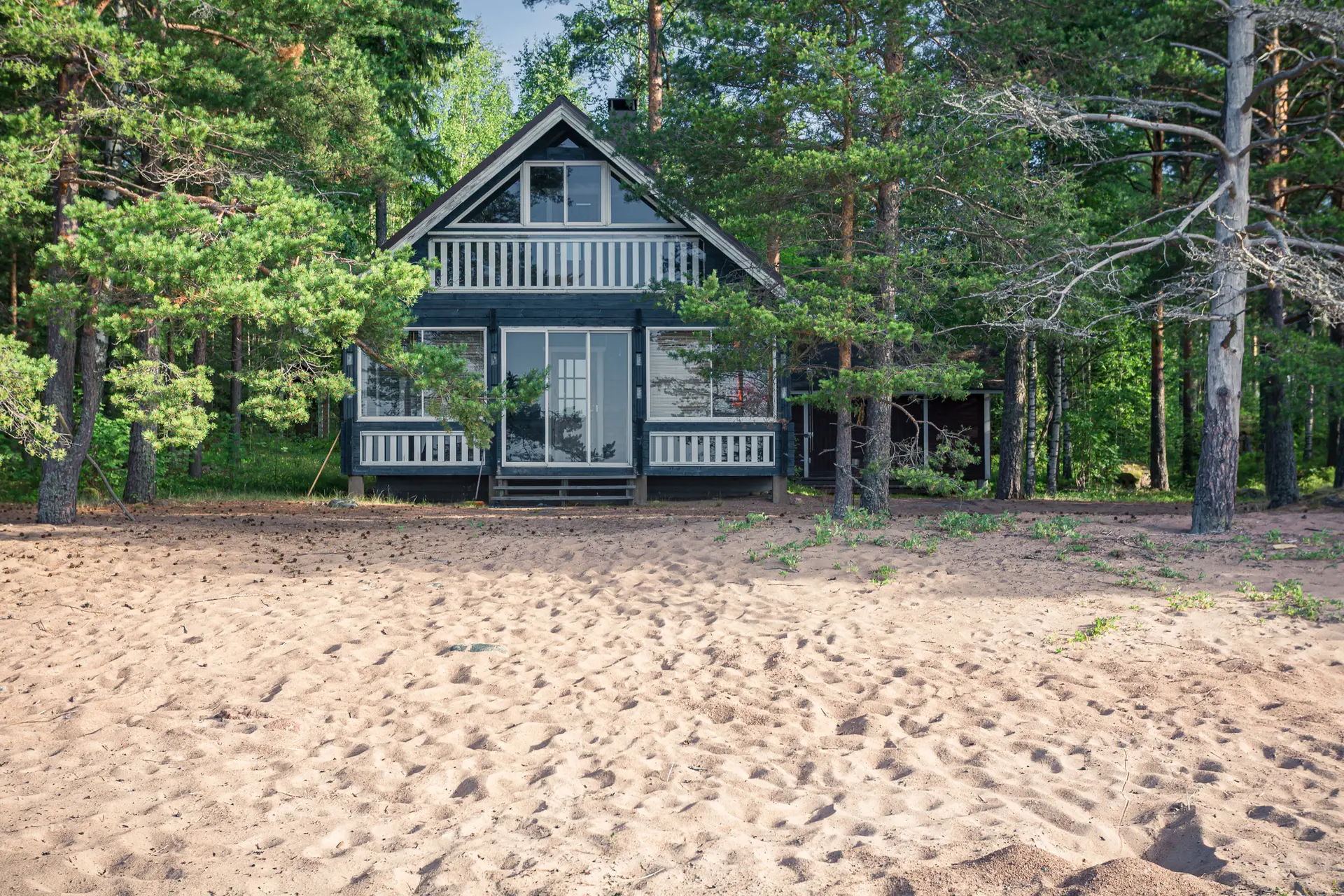 Schwarzes Ferienhaus mit weißem Balkon, umgeben von Kiefern, am sandigen Ufer in Virolahti.