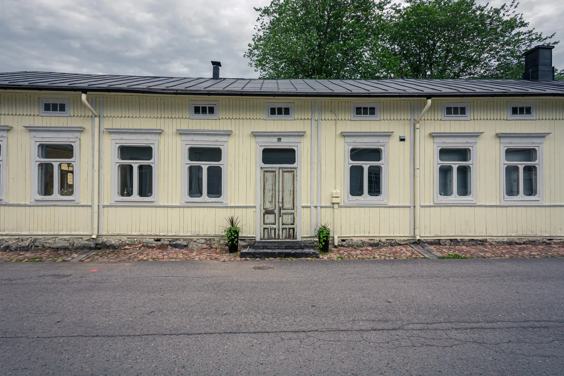 Gelbes Holzgebäude mit mehreren Fenstern und einer zentralen Eingangstür, umgeben von einem gepflasterten Weg und bepflanzten Kübeln.