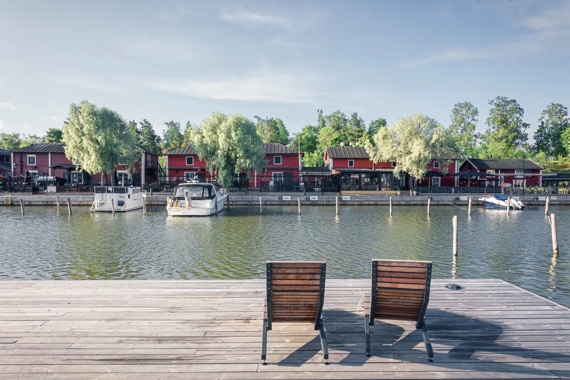 Zwei Holzliegestühle auf einem Steg mit Blick auf ein ruhiges Gewässer, umgeben von roten Häusern und grünen Bäumen.