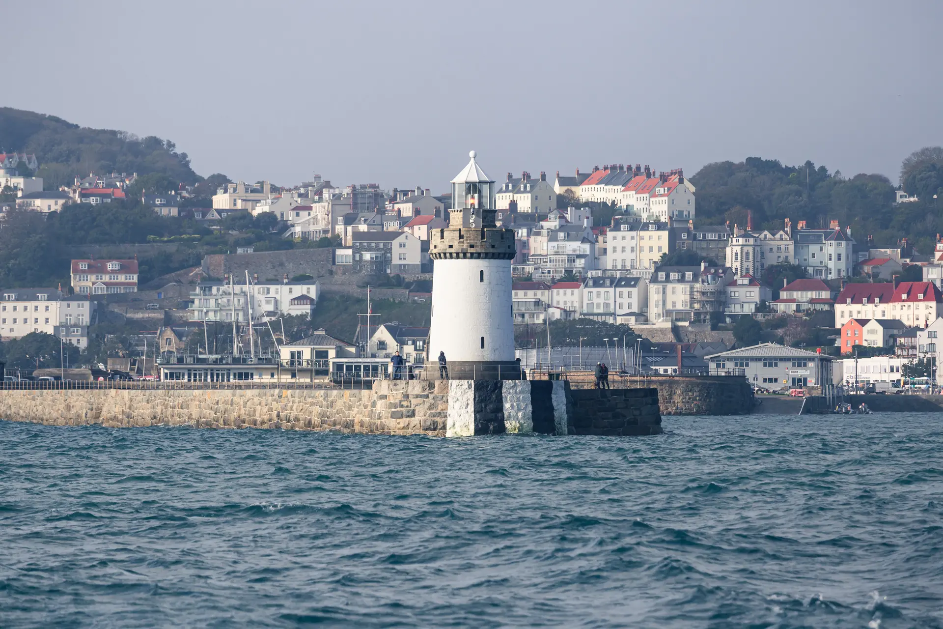 A lighthouse on a rocky pier with colorful buildings in the background, surrounded by calm blue waters.