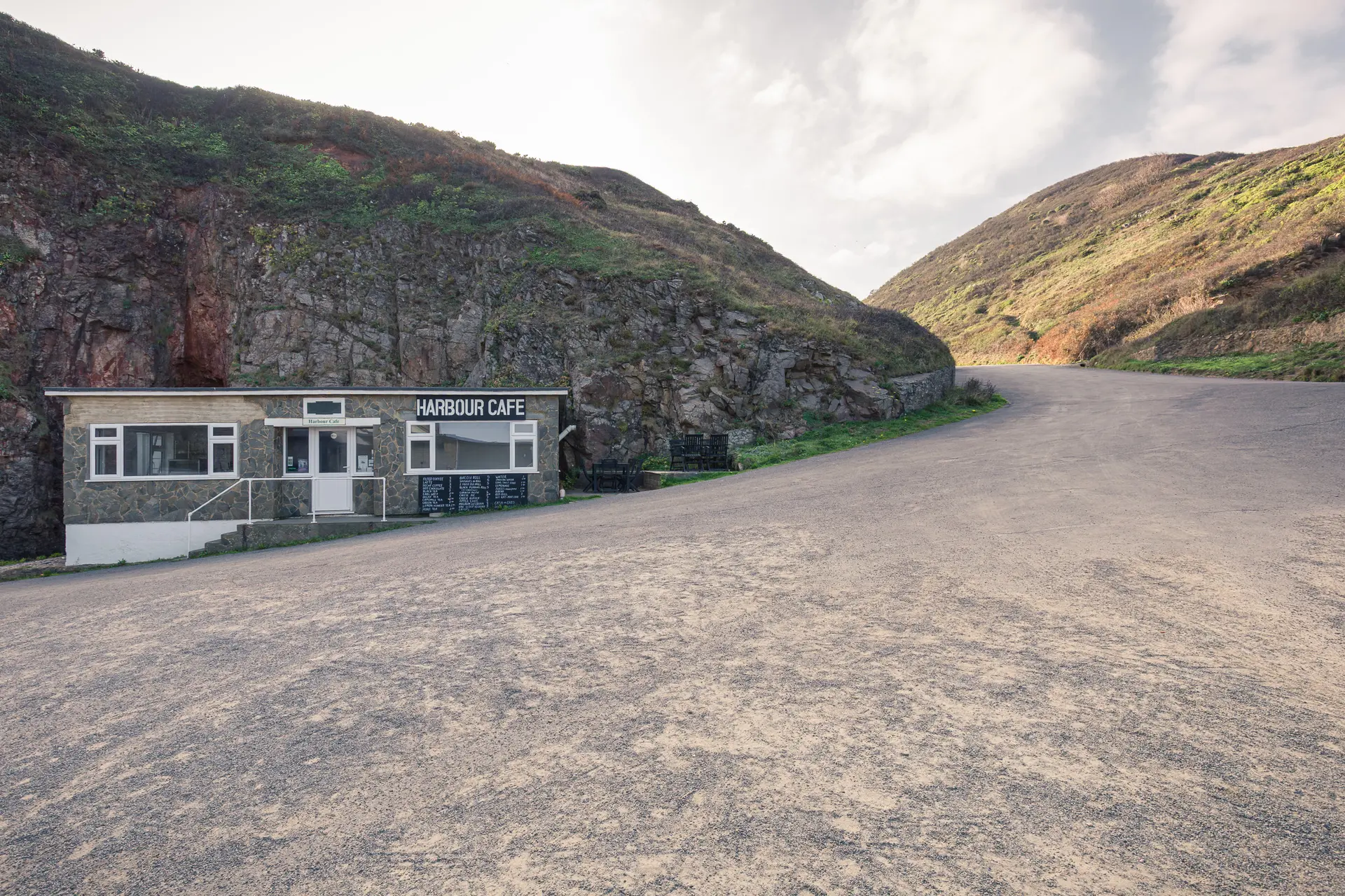 A coastal café nestled between rugged hills, with a winding road leading up to it, surrounded by green landscapes.