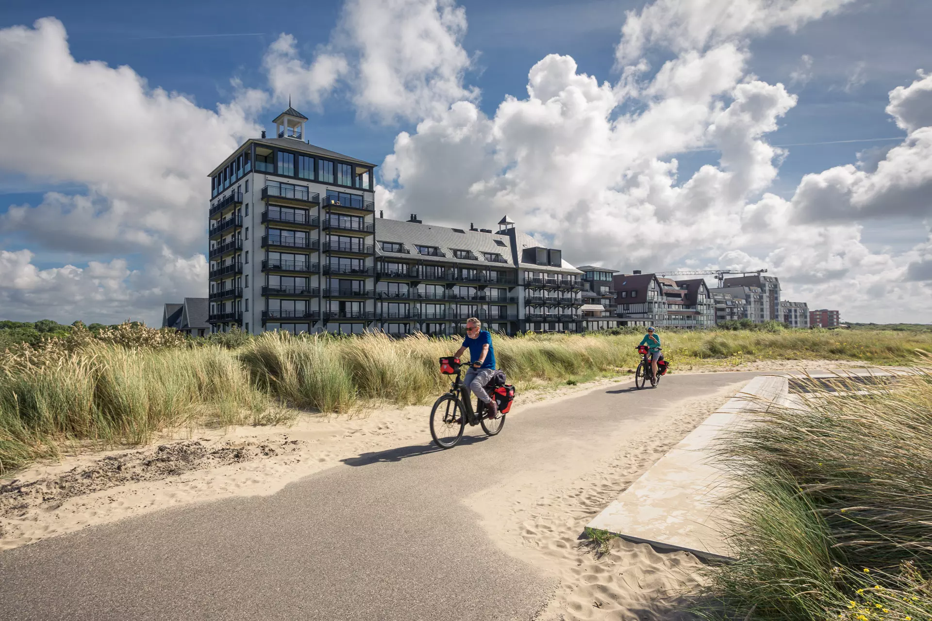 Zwei Radfahrer fahren auf einem Betonweg zwischen Sand und hohen Gräsern, mit modernen Gebäuden und blauem Himmel im Hintergrund.