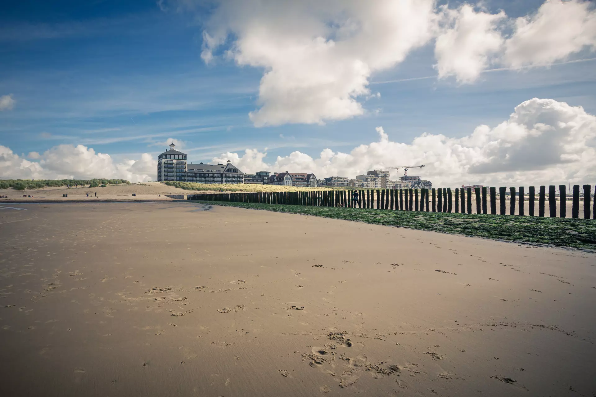 Sandy beach with wooden posts, a large building in the background, and a partly cloudy sky. People are visible in the distance.