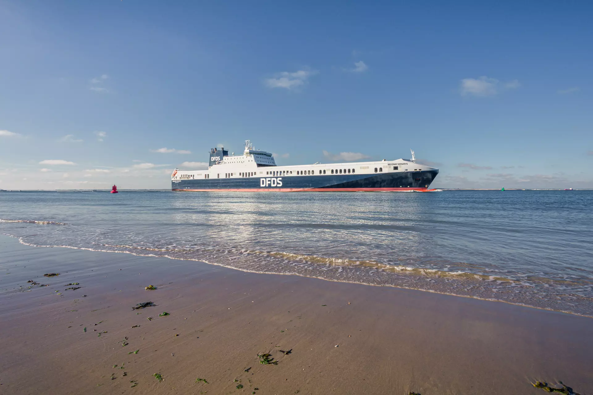 A ferry is passing on calm waters, with a clear blue sky and sandy beach in the foreground.