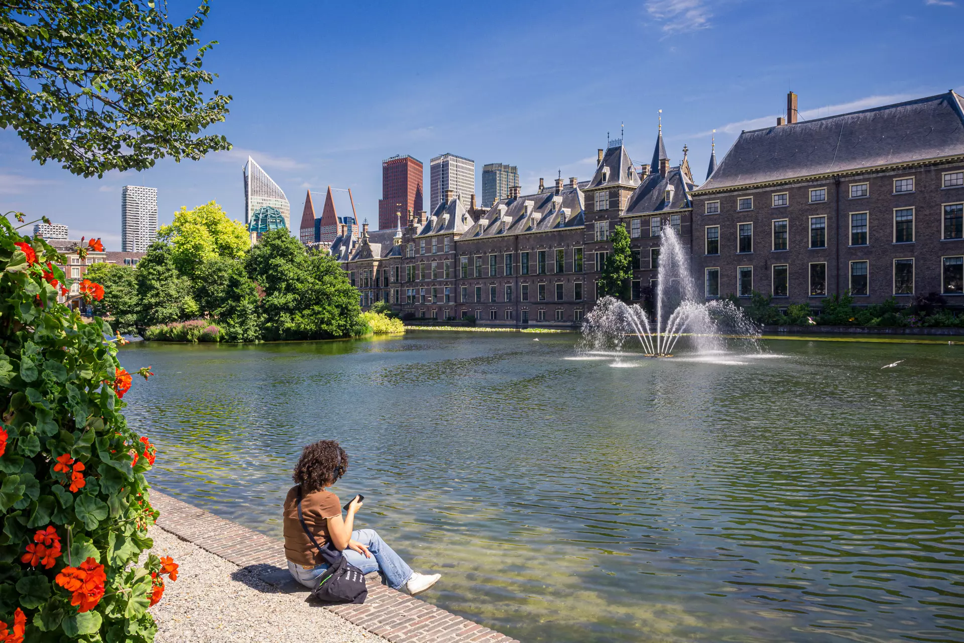 A person sits by the water's edge surrounded by flowers, facing historic buildings and a modern skyline in the background.