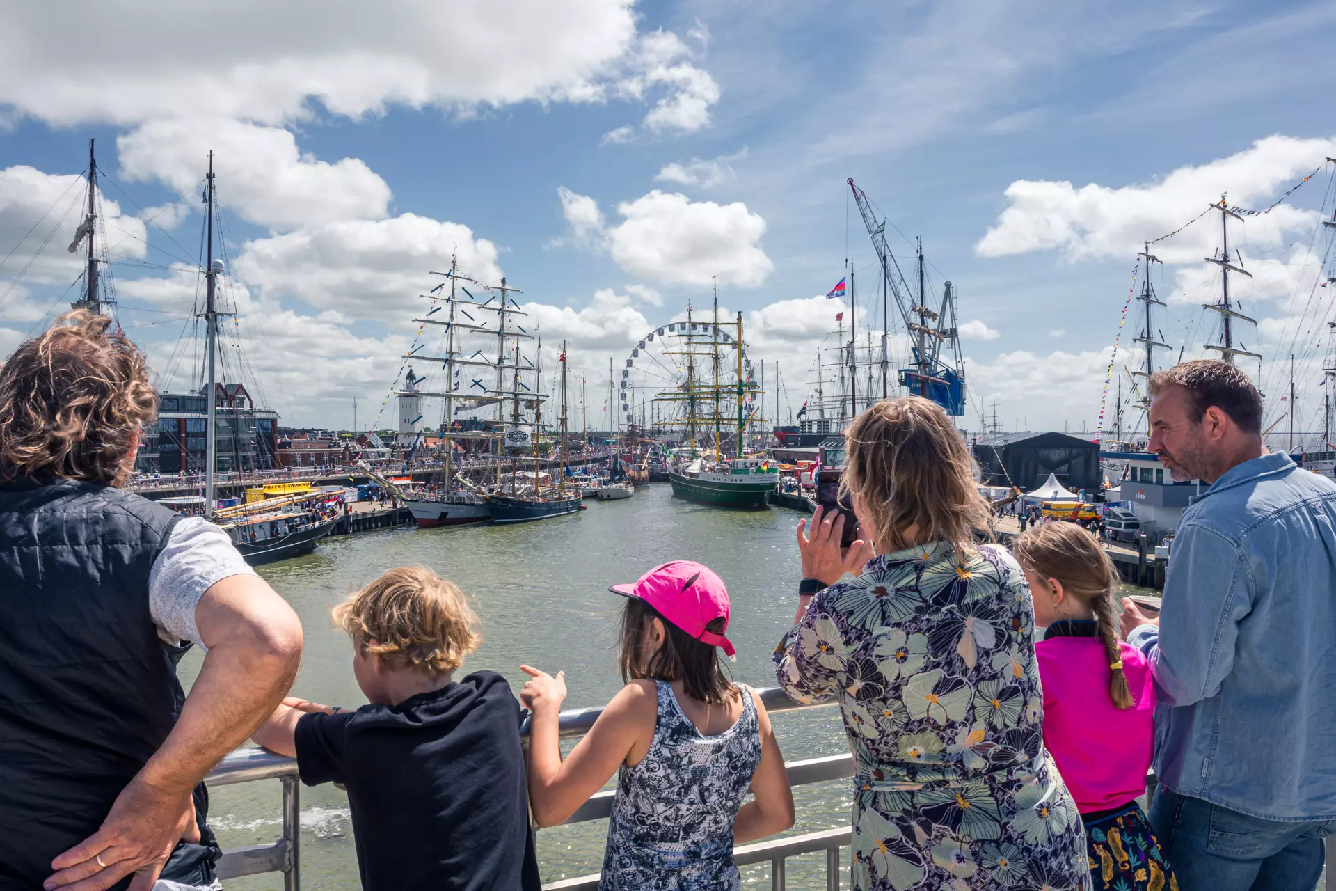 A group of people overlooks a harbor with historic ships and a Ferris wheel in the background under a blue sky.