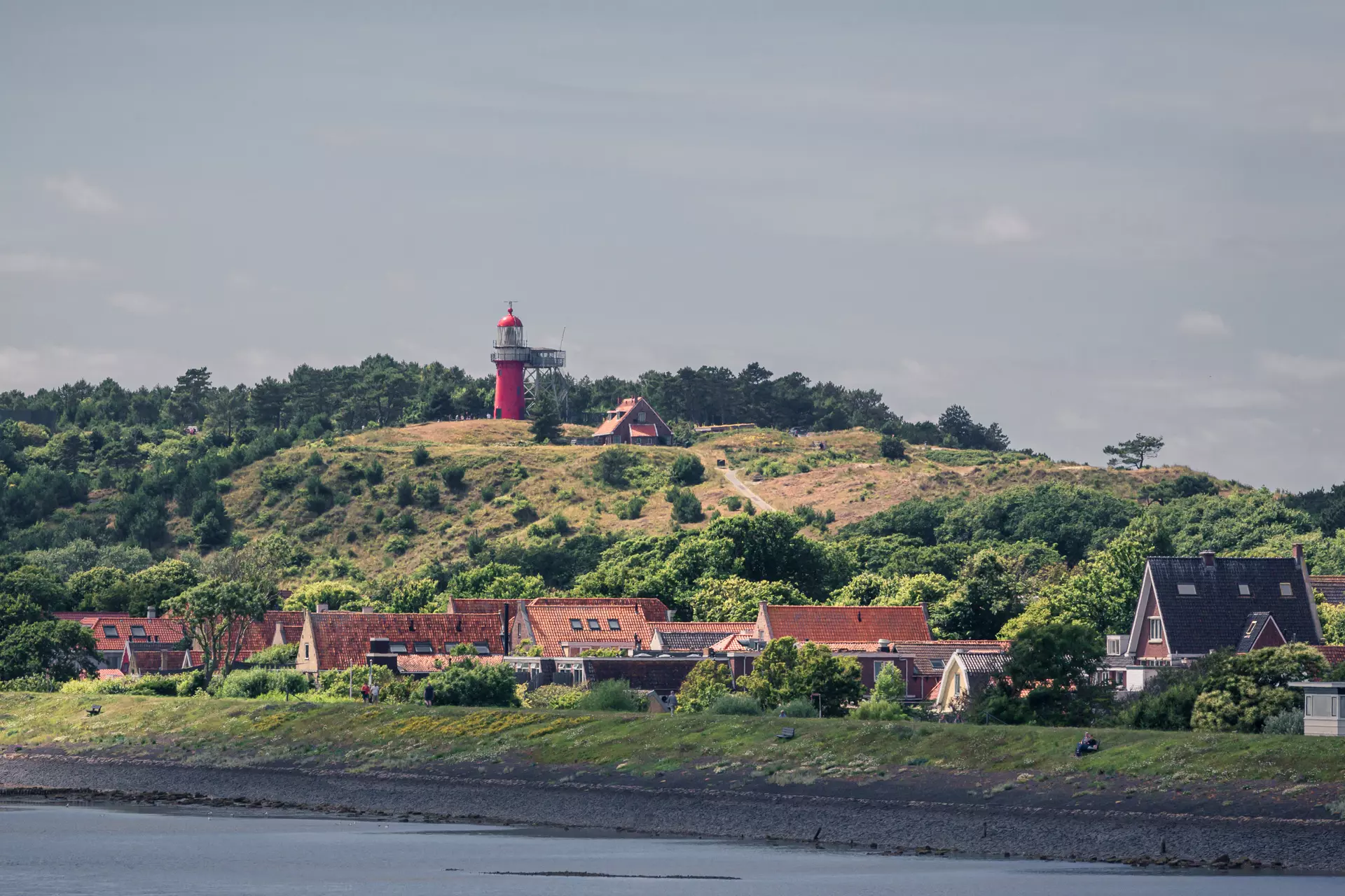 A red lighthouse sits atop a green hill overlooking colorful houses by the shore.