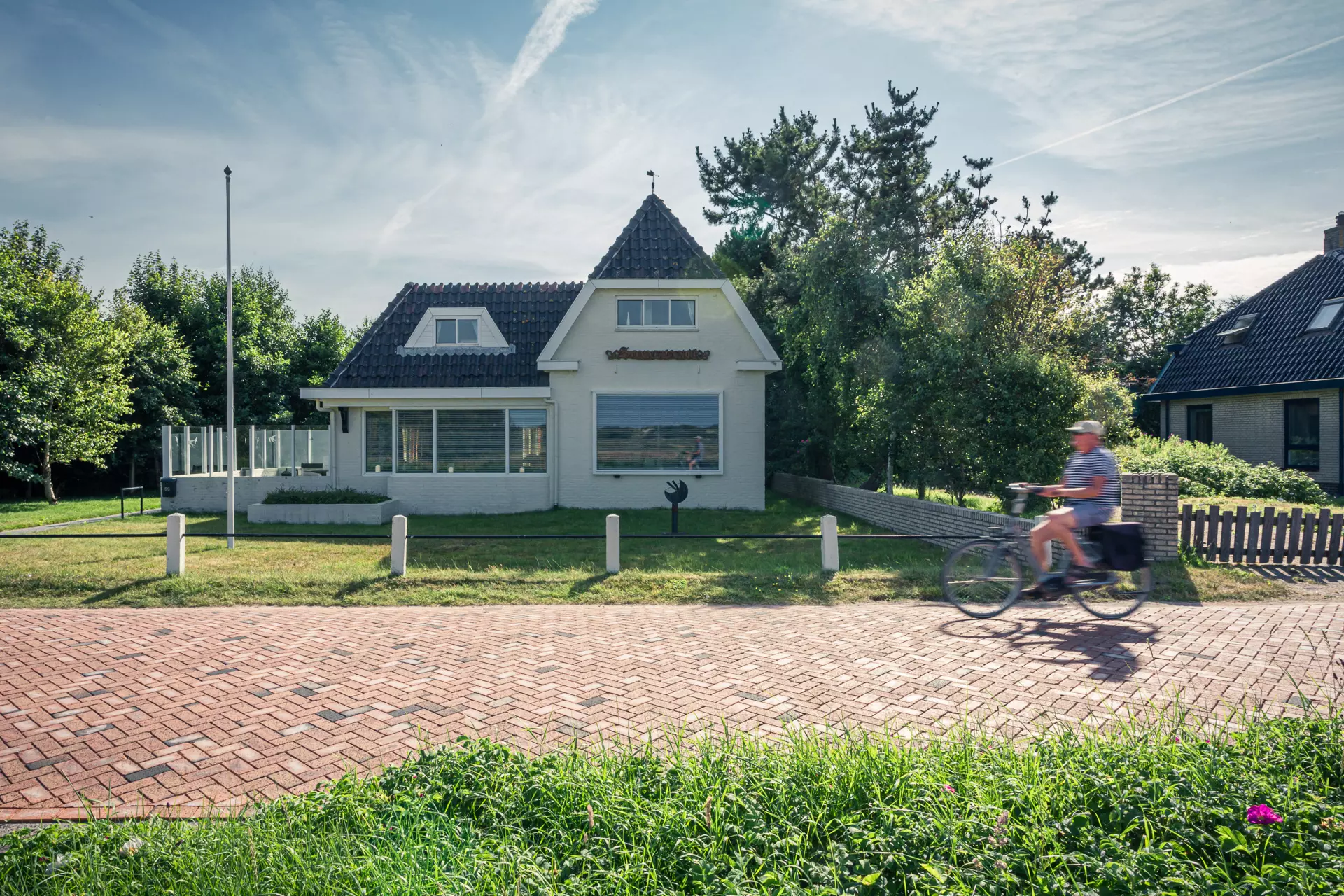 Ein Fahrradfahrer fährt an einem modernen, hellen Haus mit einem spitzen Dach und umliegendem Grün vorbei, umgeben von einem gepflasterten Weg.