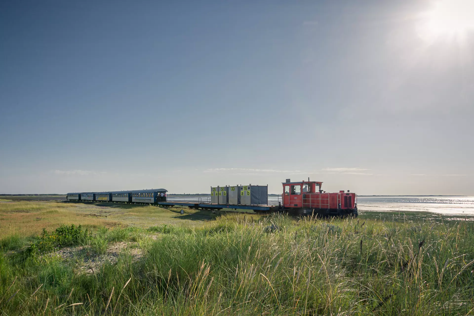 Ein kleiner roter Zug fährt an einem grasbewachsenen Ufer entlang, im Hintergrund das Meer und ein klarer Himmel.