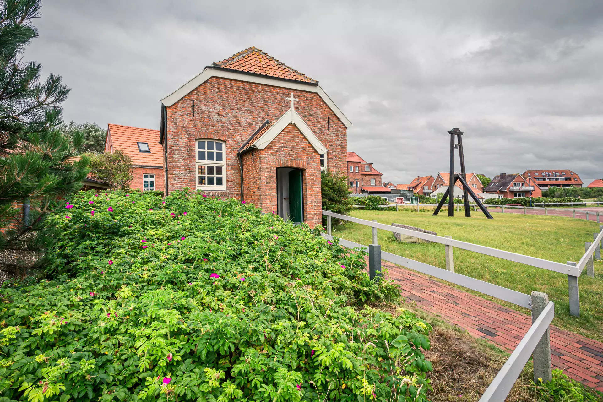 A small brick building with a gabled roof and a cross, surrounded by green plants and a small lawn.