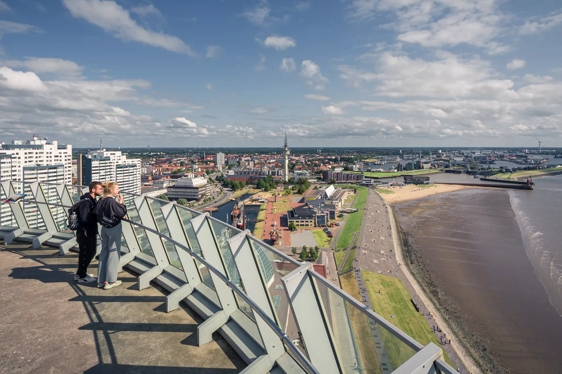 Two people stand at a railing overlooking a coastal city, surrounded by water, beaches, and clouds.