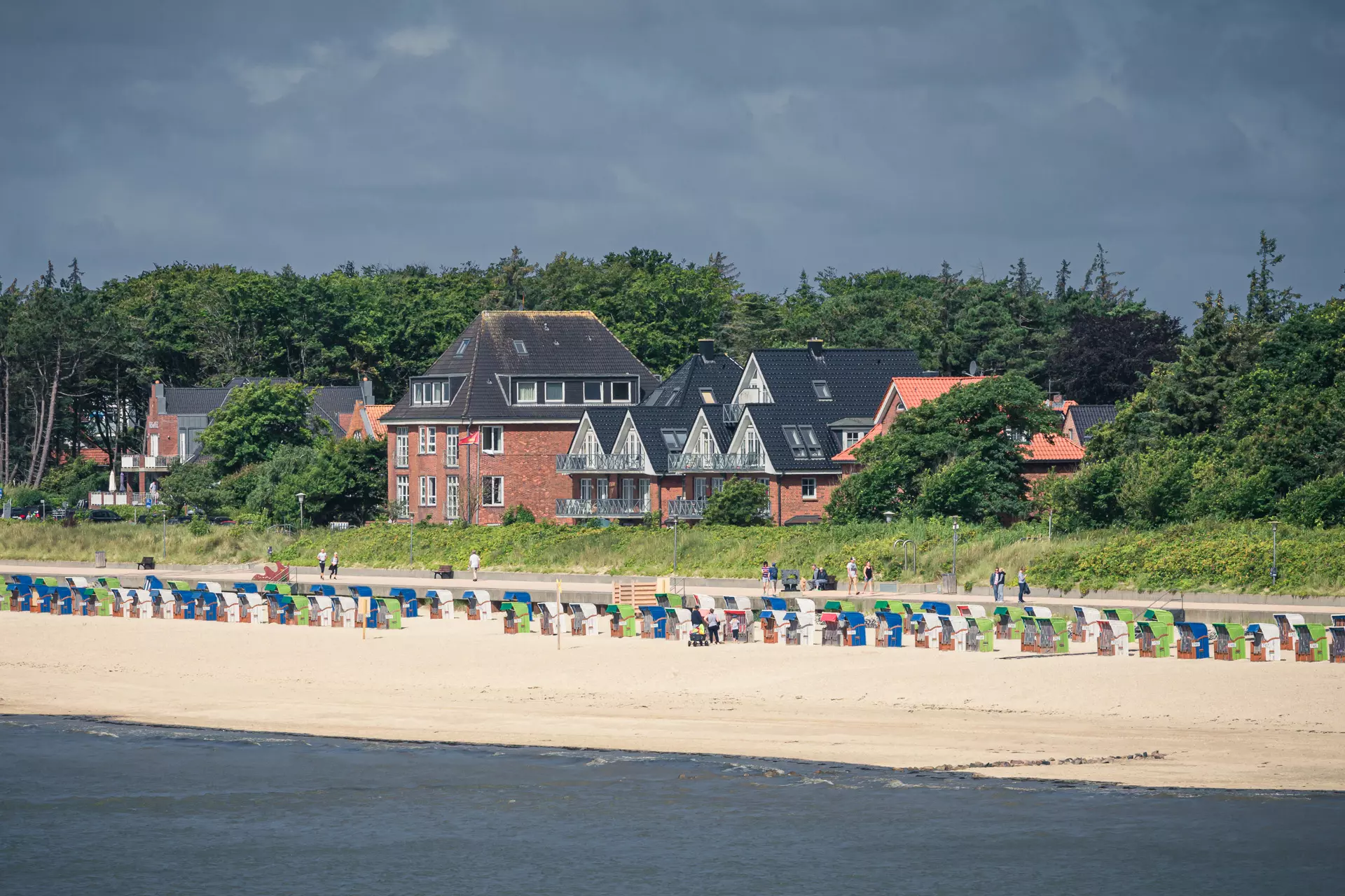 Bunte Strandkörbe stehen am Sandstrand, während im Hintergrund historische Gebäude und grüne Bäume sichtbar sind.