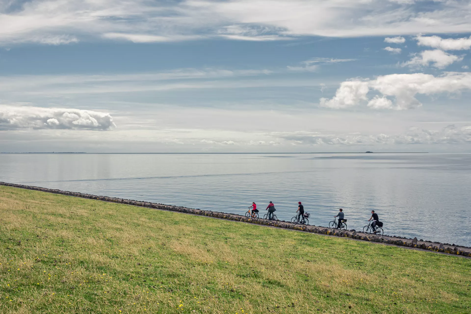 Radfahrer entlang eines Deichs mit Wasser auf der einen Seite und grüner Wiese auf der anderen. Weite Wolkenformationen am Himmel.